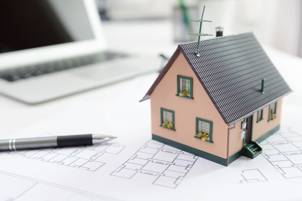 A miniature model of a house is placed on architectural blueprints, symbolizing home investment opportunities, with a pen and a laptop in the background.