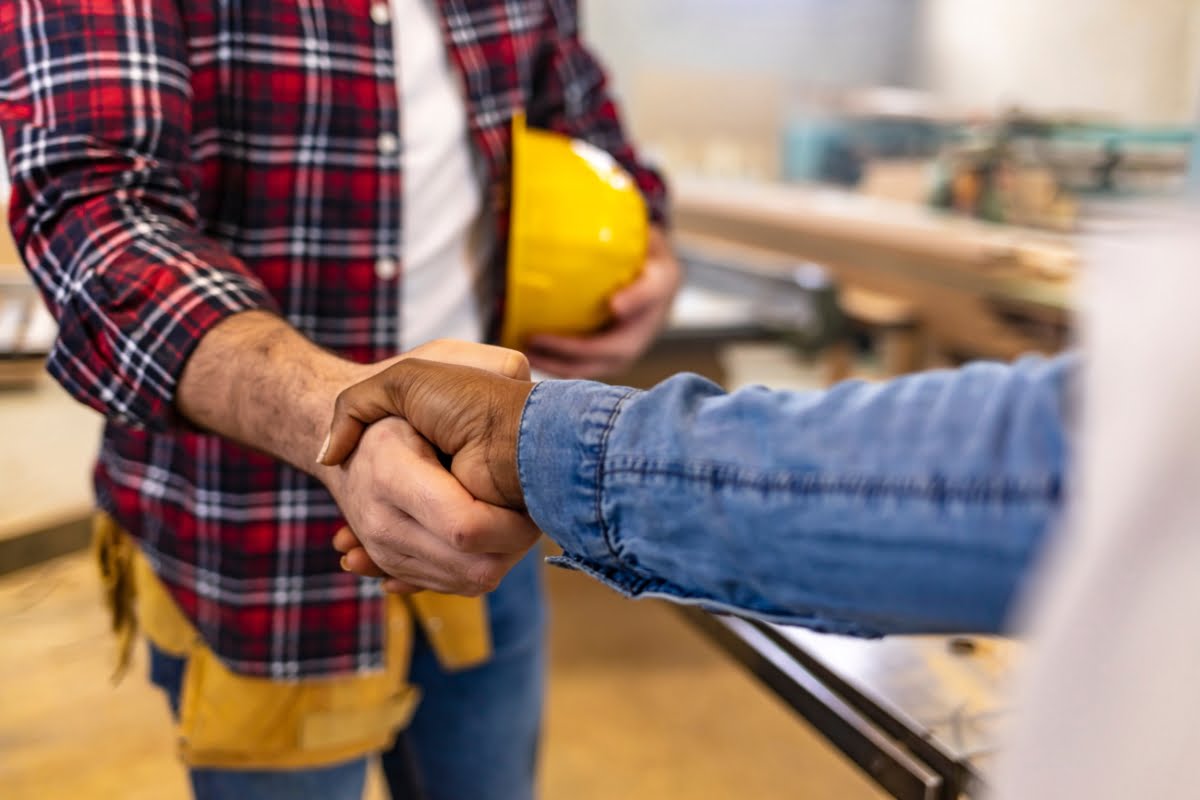 Two people shaking hands. One person, wearing a plaid shirt, holds a yellow hard hat with their other hand. Only their torsos and arms are visible, signifying a home investment partnership in progress.