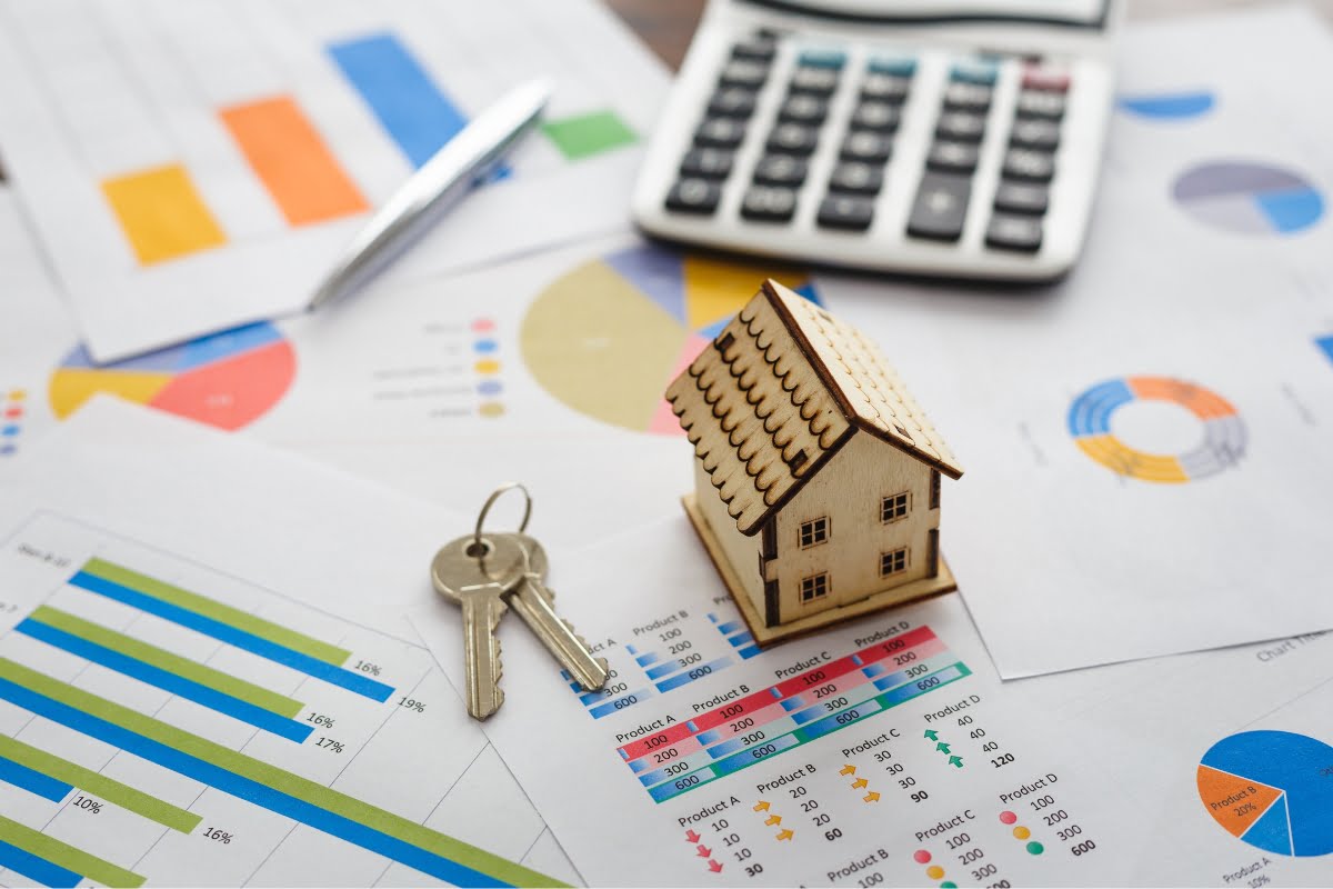 A small wooden house model and a set of keys sit on financial charts and graphs, symbolizing home investment, next to a calculator and a pen.