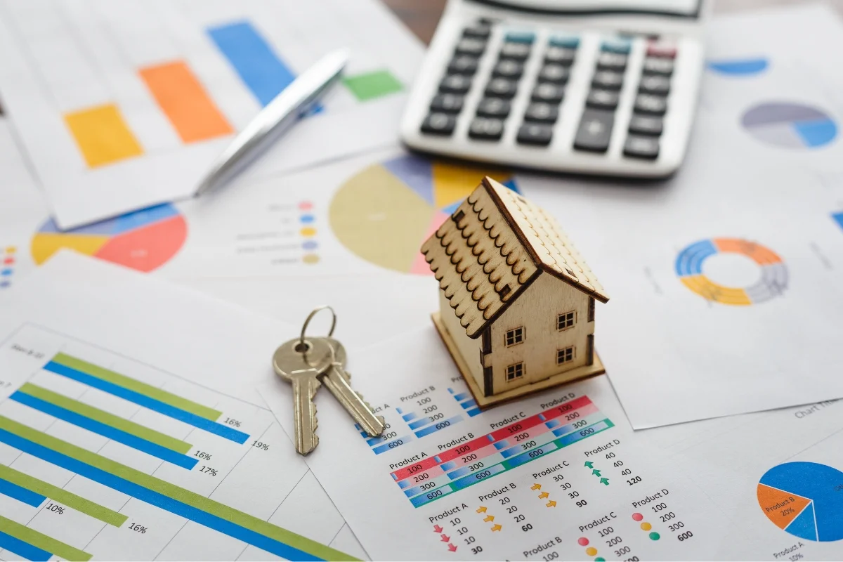 Maximize Returns: Mastering Home Investment for Modern Remodeling 9 A small wooden house model and a set of keys sit on financial charts and graphs, symbolizing home investment, next to a calculator and a pen.