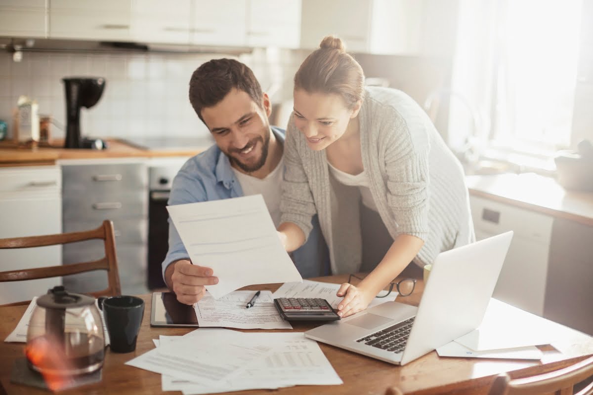 A man and a woman are reviewing documents together at a kitchen table, with a laptop, calculator, and various papers spread out in front of them. They're meticulously planning their home investment.