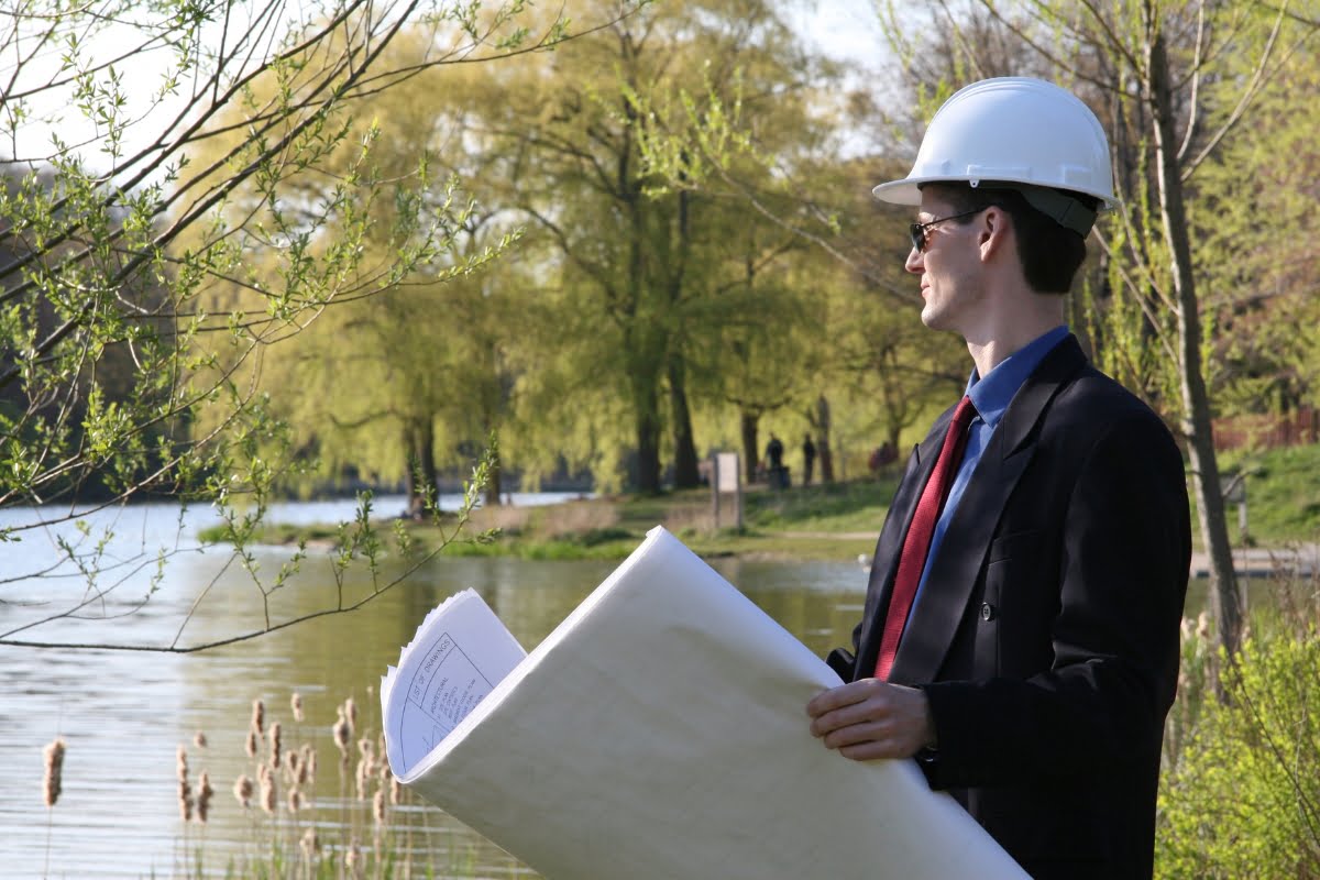 A person in a suit and hard hat stands beside a lake, holding blueprints. The serene setting contrasts with the bustling world of urban planning, while trees and water are visible in the background.