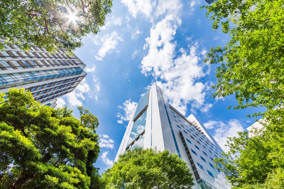 A low-angle view of modern skyscrapers, a testament to urban planning, surrounded by lush green trees under a bright, partly cloudy sky.