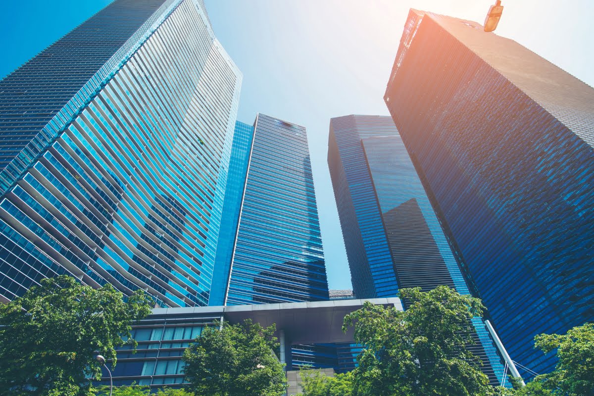 View looking up at four modern skyscrapers with glass exteriors in a city, exemplifying innovative urban planning, framed by sunlight and a few green trees at the base.