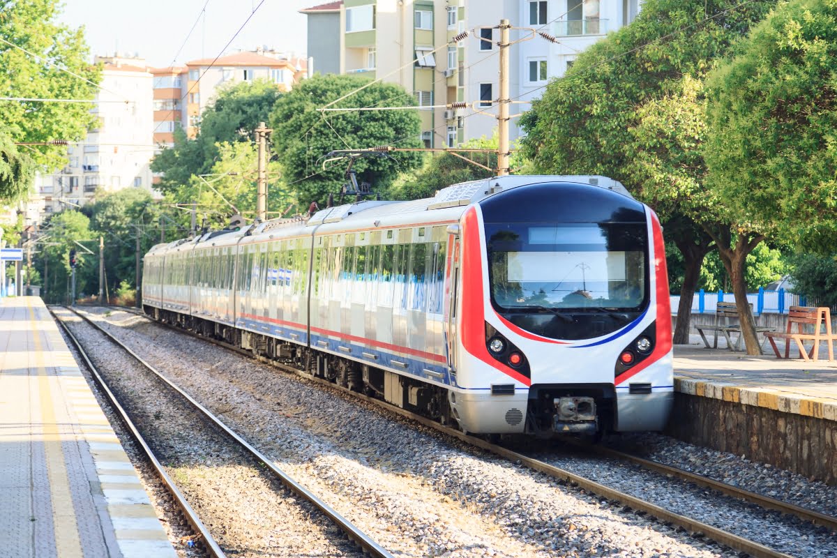 A modern train, featuring a white and red exterior, travels on tracks through a suburban area thoughtfully designed with apartments and greenery in the background, showcasing excellent urban planning.