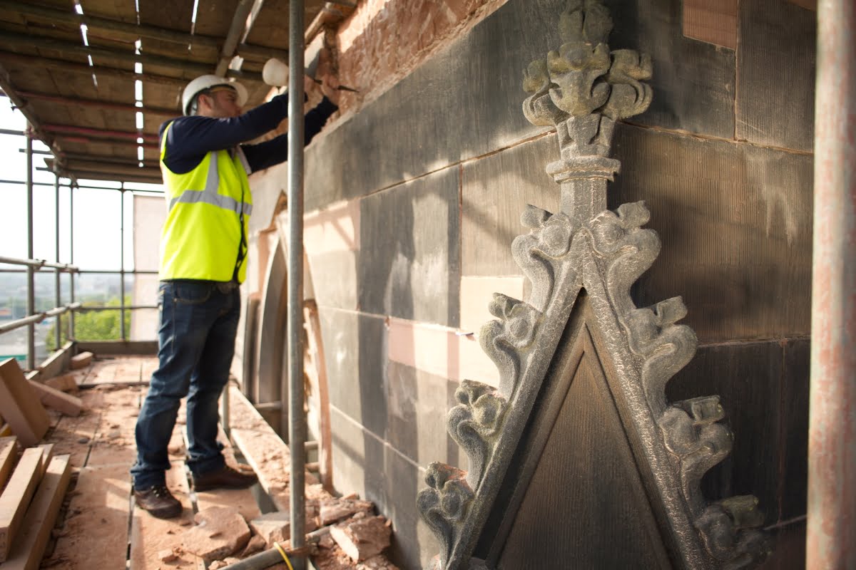 Worker in high-visibility vest and helmet conducts restoration work on an ornate building facade using scaffolding for support, contributing to comprehensive urban planning efforts.