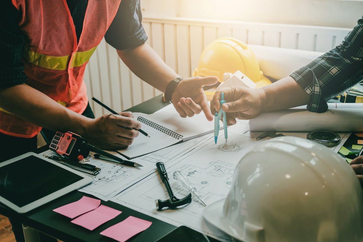 Two construction workers discussing blueprints at a table with a variety of tools, a notebook, a tablet, and construction helmets, delve into the details of urban planning strategies.