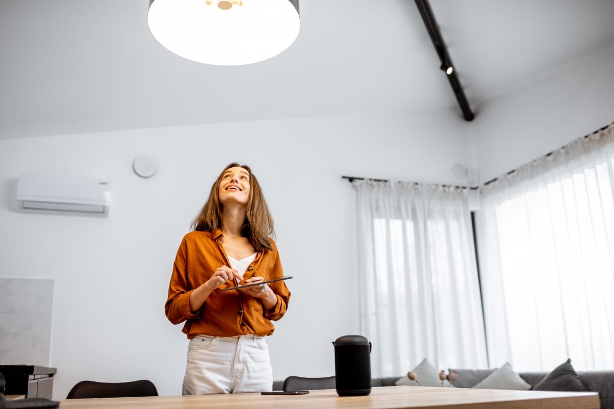 A person in an orange shirt stands with a tablet in a well-lit room, looking up and smiling, envisioning possibilities for energy-efficient remodeling.