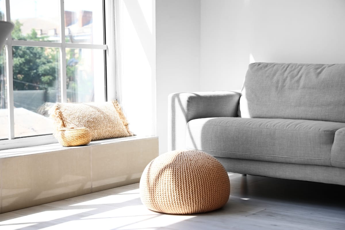 A sunlit living room showcases energy-efficient remodeling, featuring a gray sofa, a woven pouf, and a window seat adorned with a cushion and small basket.