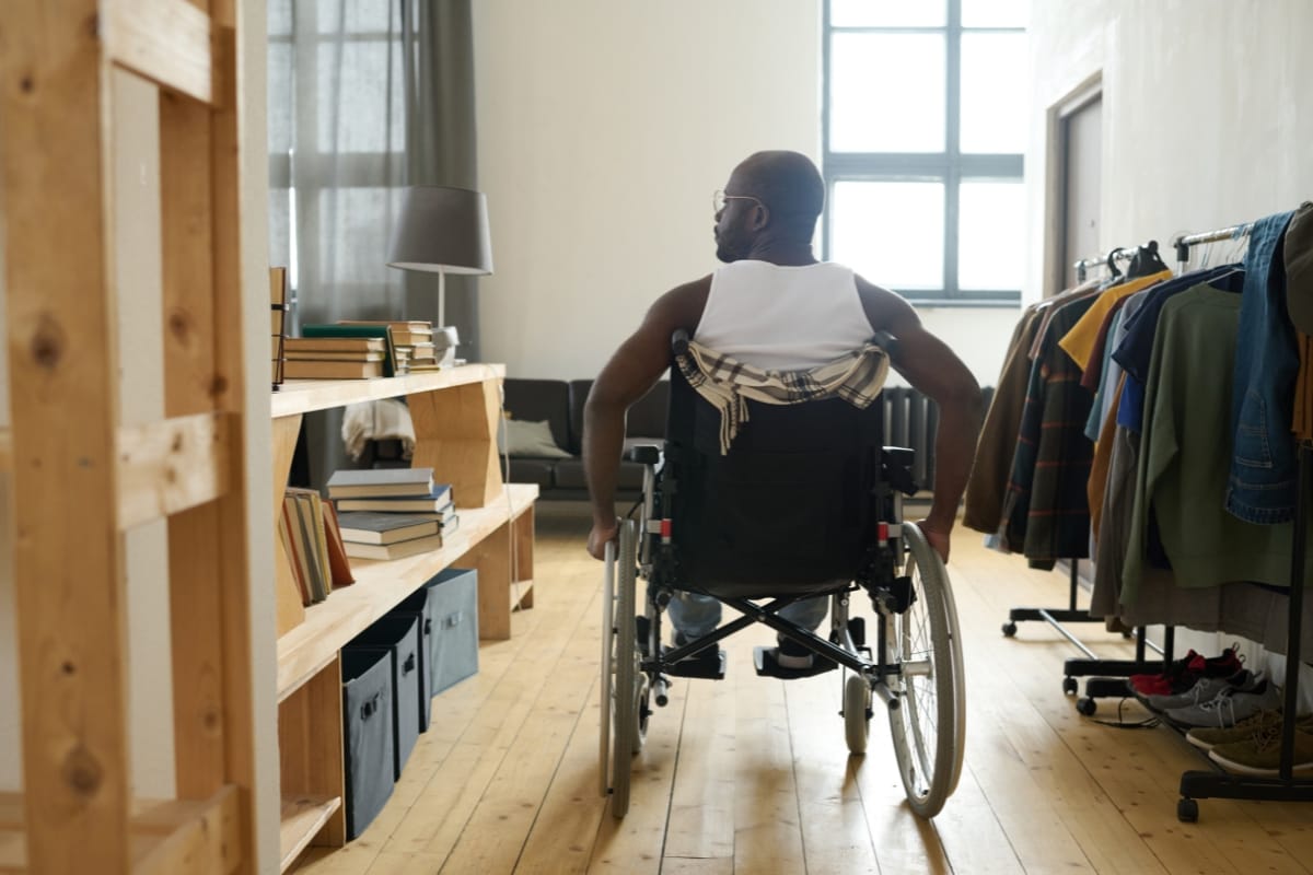 A person in a wheelchair moves effortlessly through a room designed for home accessibility, featuring wooden floors, shelves lined with books, and a clothing rack.