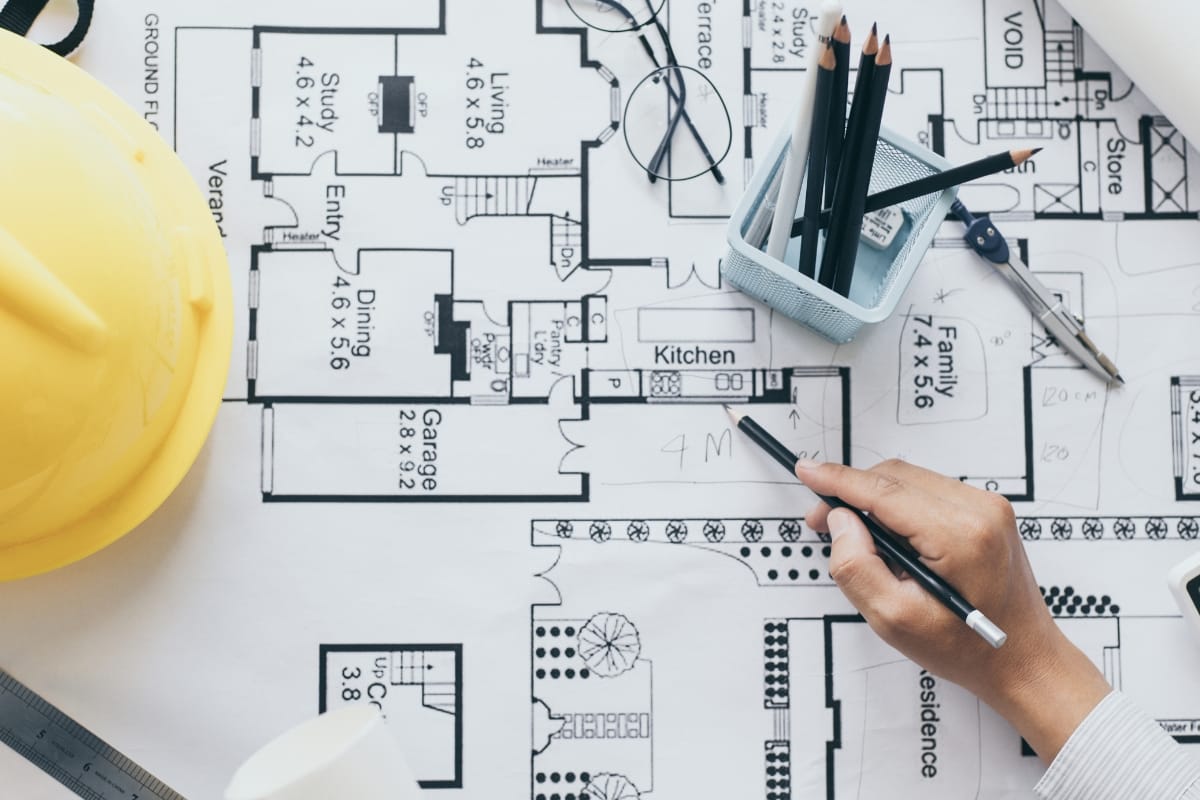 A person is sketching on architectural floor plans with a pencil, planning a home accessibility remodeling project. Nearby are a yellow hard hat, a pair of eyeglasses, and a container with pencils and pens.