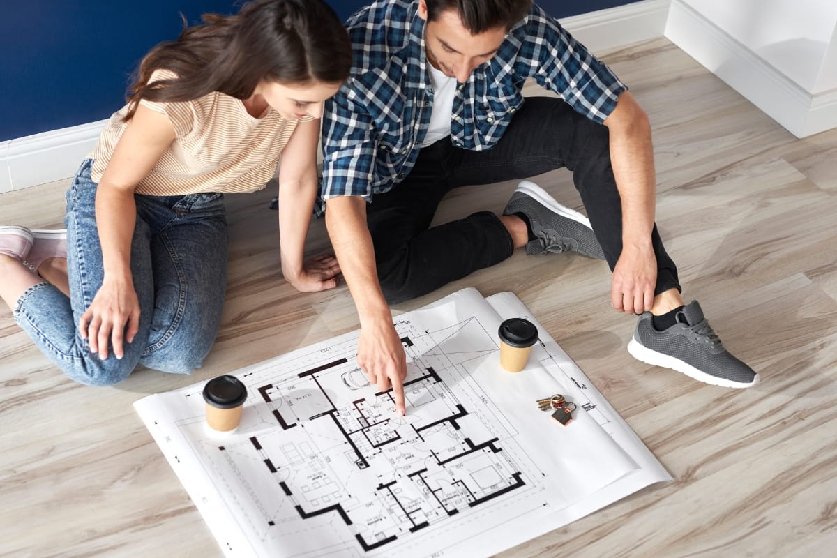 Two people sitting on a wooden floor are immersed in reviewing architectural plans for home accessibility. Their coffee cups sit nearby, fueling their creative remodeling ideas.