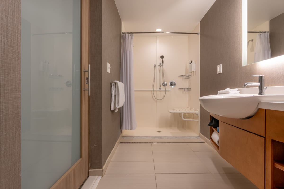 A modern bathroom featuring a walk-in shower with a sliding glass door, sink with towel, and vanity mirror. The design, perfect for home accessibility remodeling, showcases brown walls complemented by beige floor tiles.