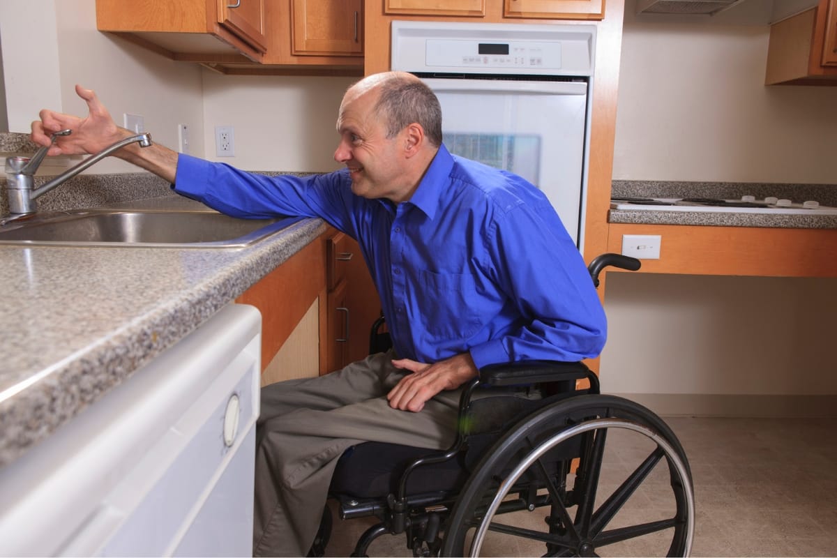 A person in a wheelchair reaches for the faucet in a kitchen thoughtfully remodeled with lower counters, showcasing smart home accessibility design.