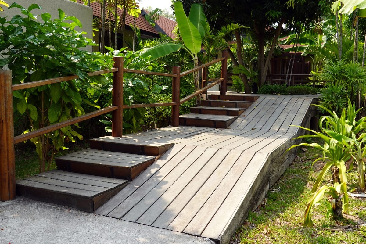 Wooden ramp and stairs with a sturdy railing provide seamless home accessibility, all surrounded by lush greenery in a serene garden setting.