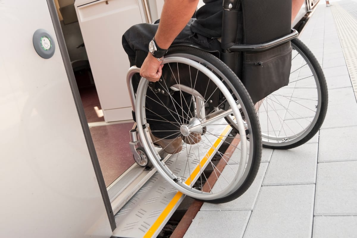 A person in a wheelchair, having recently benefited from home accessibility remodeling, uses an access ramp to board a train at the station.