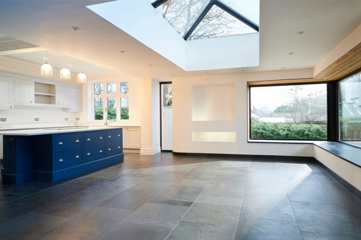 This spacious modern kitchen in an accessory dwelling unit features a blue island, skylight, large windows, and slate flooring. White cabinets line the wall, with pendant lights elegantly hanging above the island.