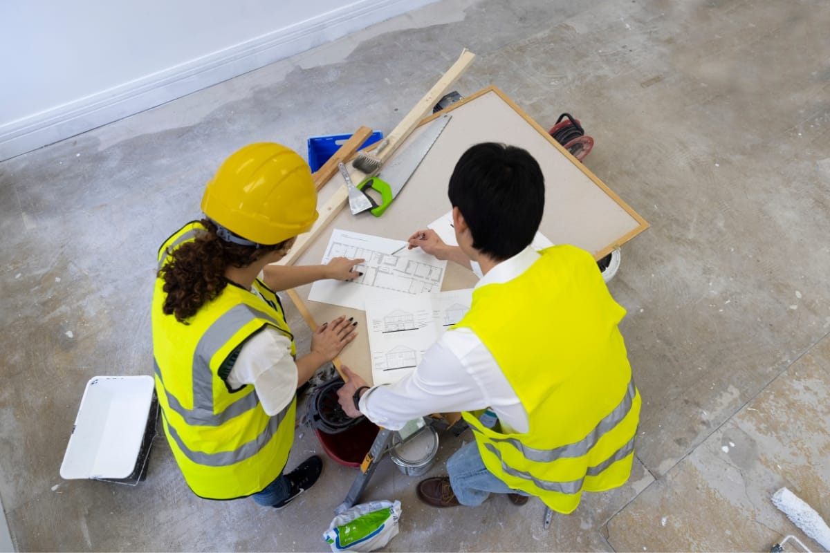 Two construction workers wearing yellow vests and hard hats review blueprints for an accessory dwelling unit on a table in a room with unfinished flooring.