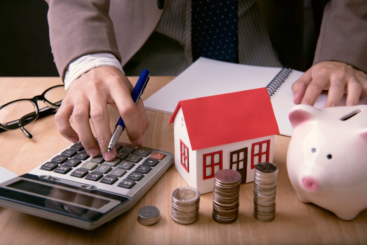 A person uses a calculator beside a model of an accessory dwelling unit, with stacked coins and a piggy bank on the desk.