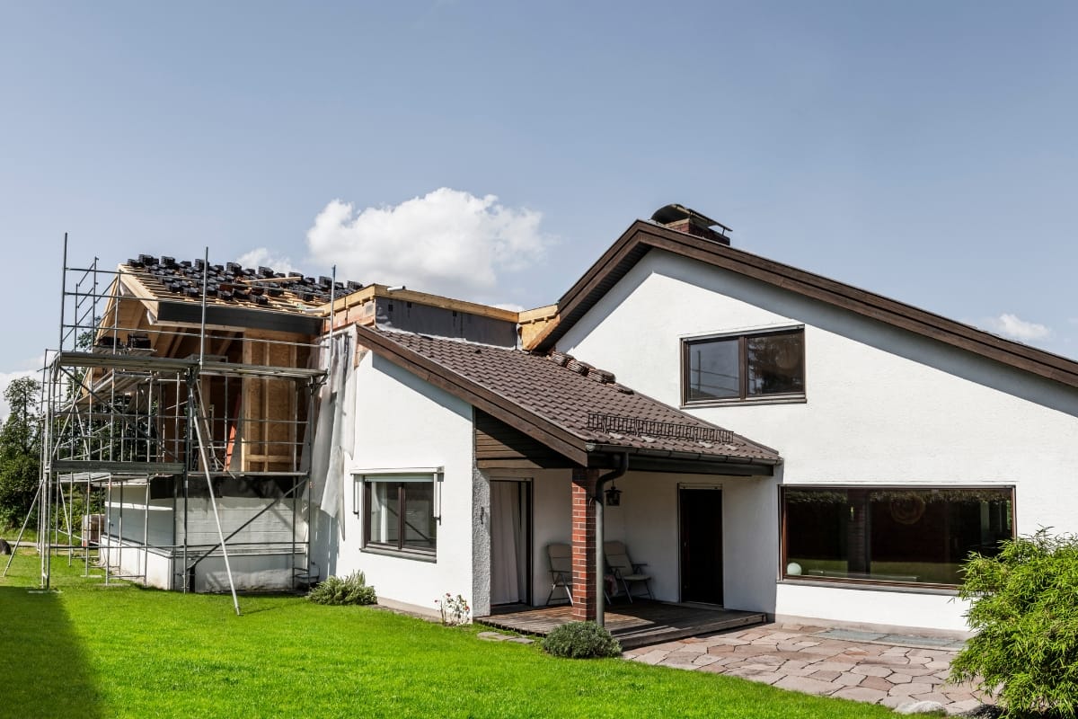 A white two-story house with a large front lawn is under renovation, possibly to include an accessory dwelling unit. Scaffolding is set up on the left side of the building.