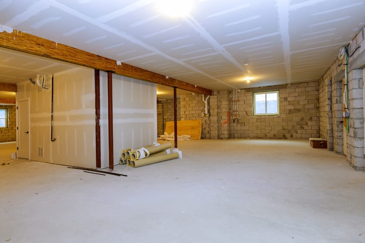 Spacious unfinished basement with concrete walls, exposed beams, and drywall. Rolls of insulation on the floor hint at potential basement finishing errors. One small window and visible wiring complete the setting.