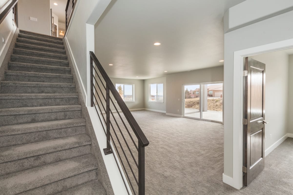 A modern, empty living room with gray carpet and large windows basks in natural light. The sleek staircase with a metal railing adds a contemporary touch, while an open doorway hints at possibilities beyond. Consider basement finishing services to extend this refined elegance below.