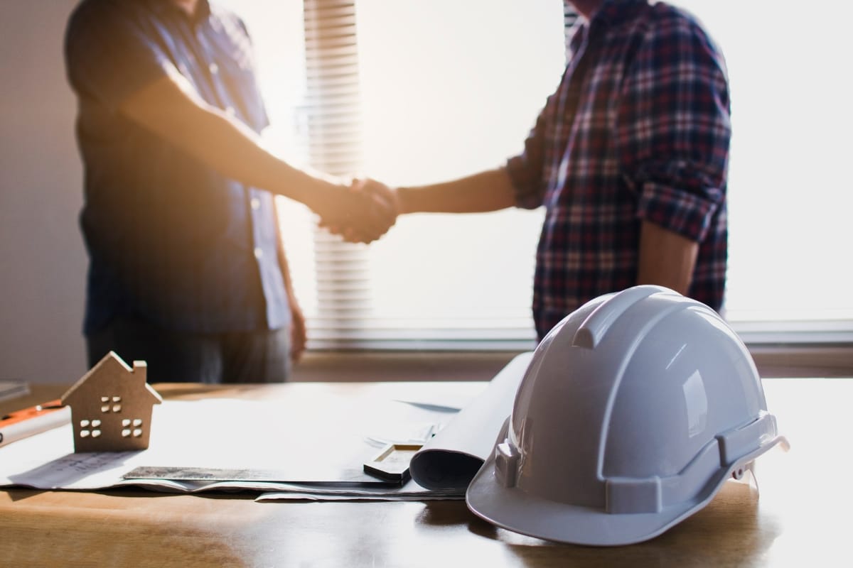 Step-by-Step Remodeling Process: How to Renovate Your Roswell Home Seamlessly 3 Two people shake hands in an office setting, with a white hard hat, architectural plans, and a small house model on the desk—signaling the start of the remodeling process.