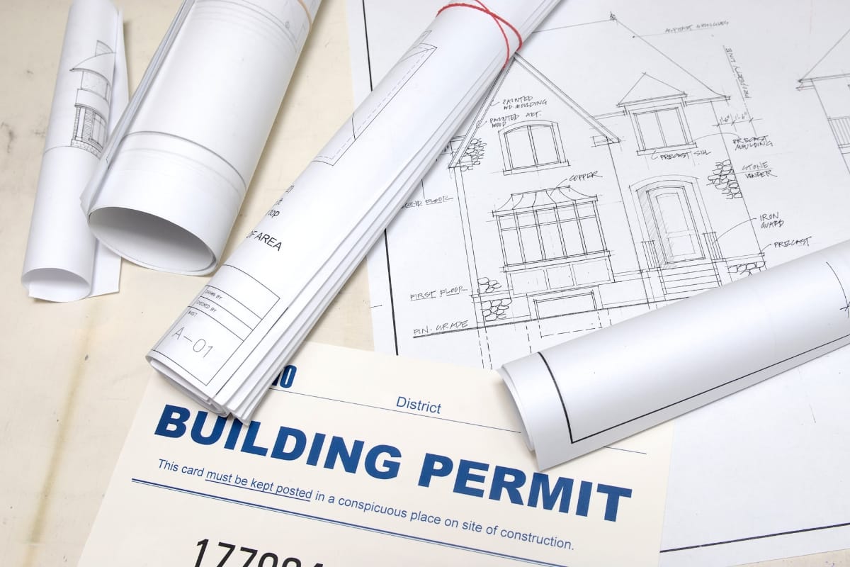 Step-by-Step Remodeling Process: How to Renovate Your Roswell Home Seamlessly 5 Rolled architectural blueprints and a building permit are displayed on a table, alongside a detailed drawing of a house exterior, illustrating the early stages of the remodeling process.