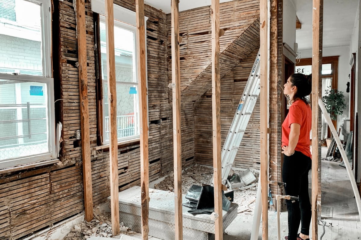 Step-by-Step Remodeling Process: How to Renovate Your Roswell Home Seamlessly 6 A woman in a red shirt stands in a partially demolished room, surrounded by exposed wall studs and debris, illustrating the remodeling process, with a ladder positioned near the window.