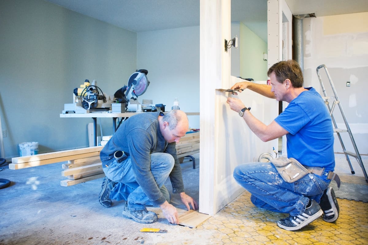 Step-by-Step Remodeling Process: How to Renovate Your Roswell Home Seamlessly 7 Two men work on a home renovation; one measures a wooden board on the floor while the other uses a tool on a door frame, highlighting the remodeling process amid scattered construction materials.