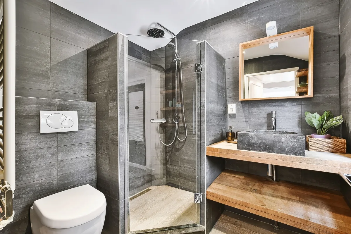 Modern bathroom remodeling with dark gray tiles, a glass-enclosed corner shower, wall-mounted toilet, and wooden vanity featuring a stone sink and potted plant.