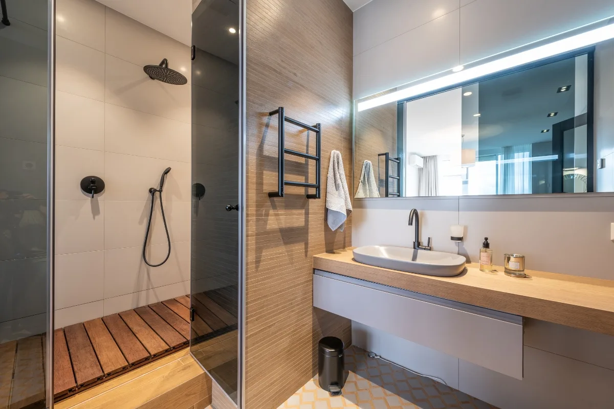 Modern bathroom remodeling features a walk-in shower with a rain showerhead, wooden floor slats, wall-mounted towel rack, large mirror, and countertop sink with toiletries.