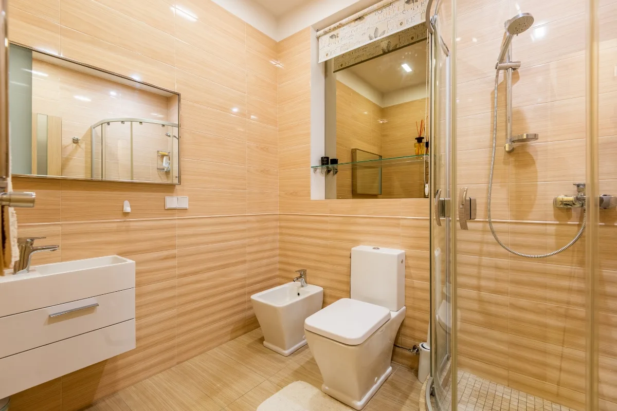 Modern bathroom with tan tiled walls, featuring a sink, bidet, toilet, glass-enclosed shower, and large mirror above the sink and shelf—an ideal showcase for stylish bathroom remodeling.