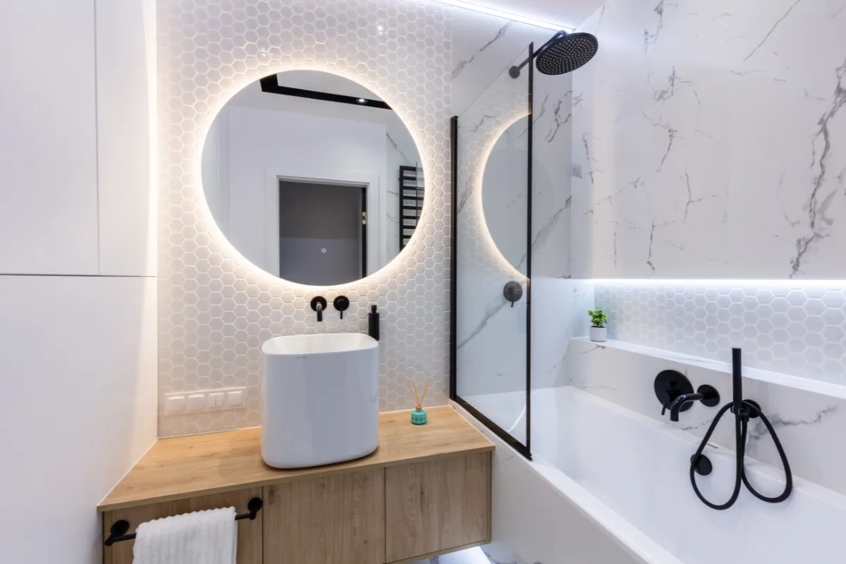 Modern bathroom remodeling features a round backlit mirror, white vessel sink on a wooden vanity, sleek black fixtures, and a bathtub with marble-patterned walls.