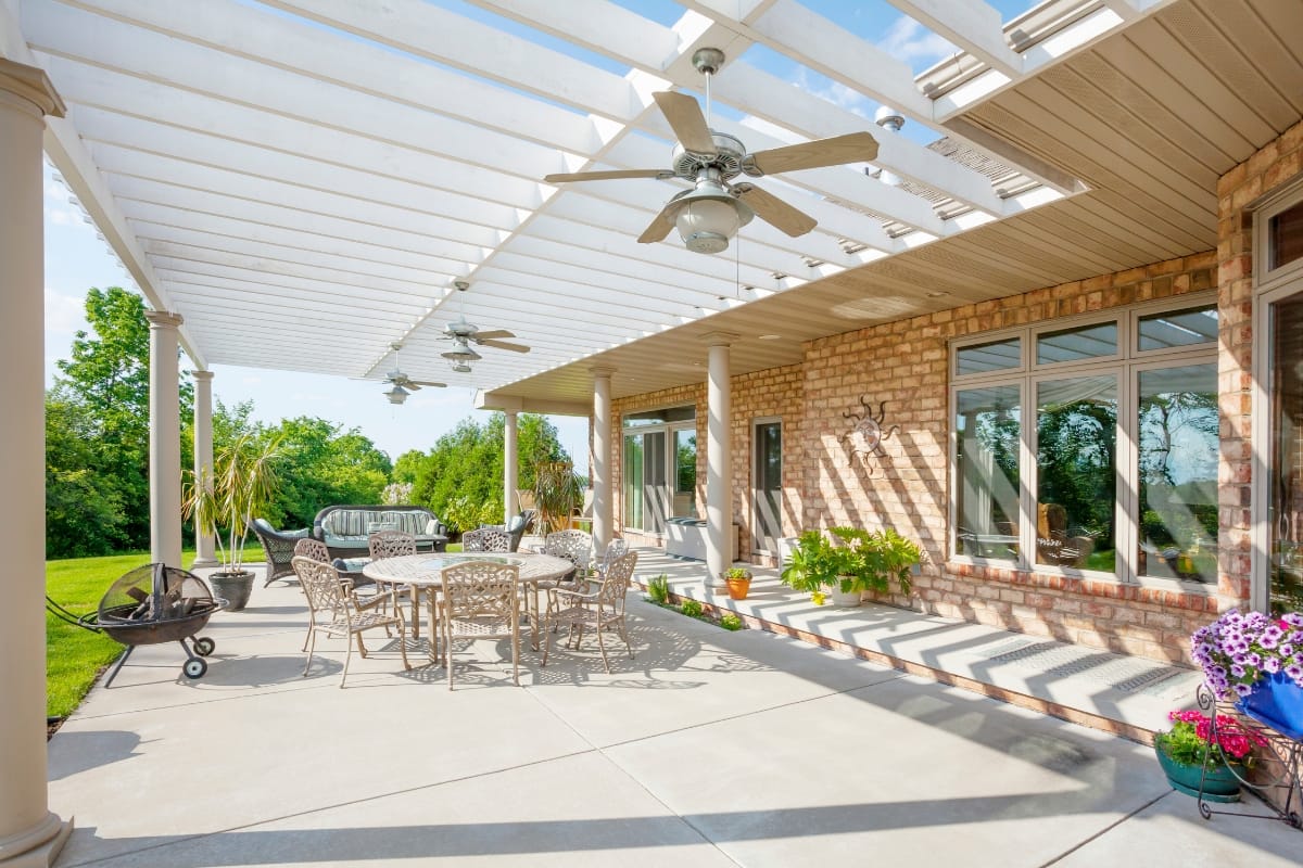 Spacious outdoor patio with pergola, ceiling fans, outdoor dining set, potted plants, and brick exterior, overlooking a green lawn and trees.