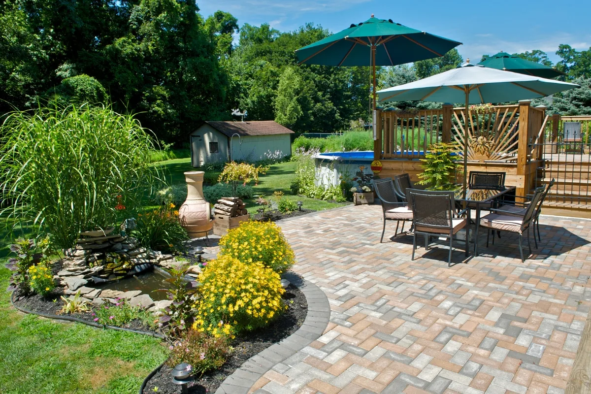 A backyard patio with paver flooring, outdoor dining set under umbrellas, a small pond with plants, and a shed in the grassy area near trees—perfect for relaxing and enjoying beautiful outdoor patios.