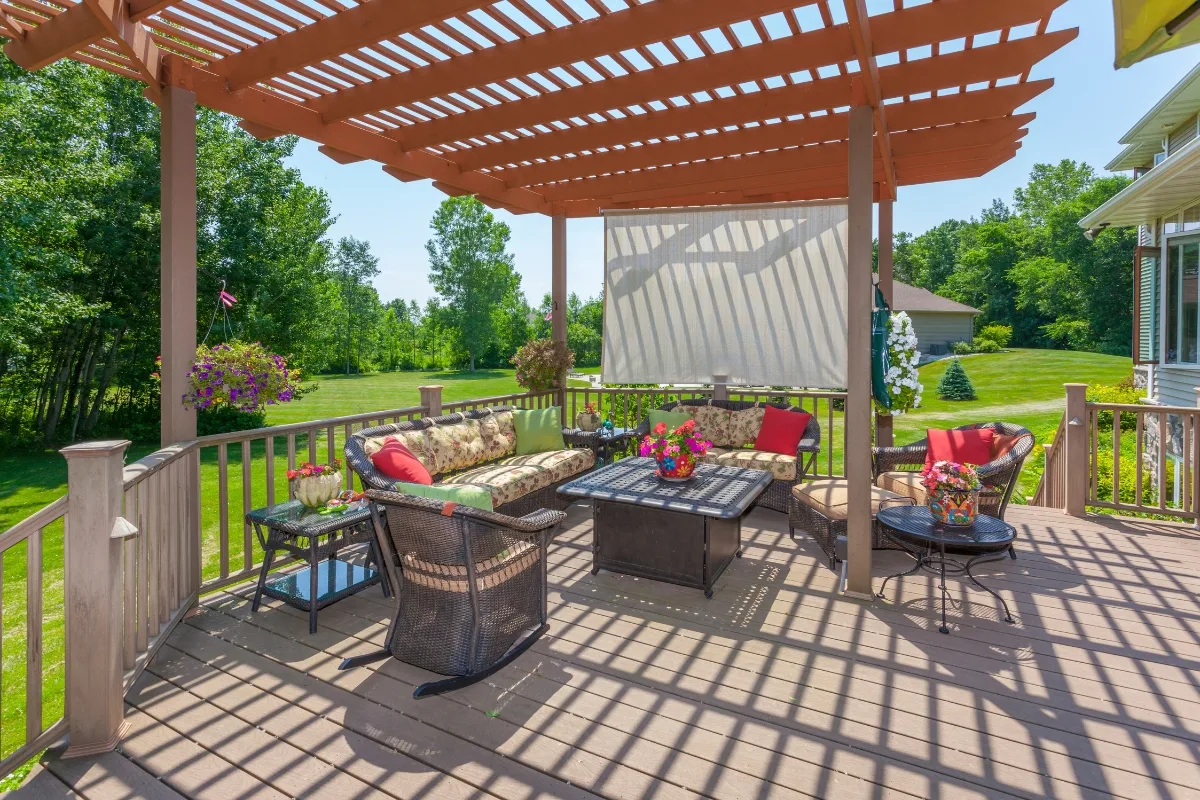 A patio with cushioned seating, a table, flowers, and a pergola casting shadows—this inviting outdoor patio overlooks a green backyard on a sunny day.