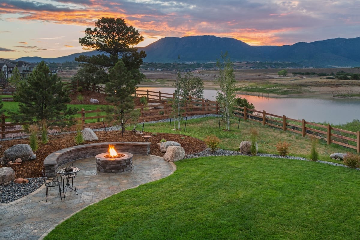 Outdoor Patios, Decks, and Fire Pits in Sandy Plains: Expert Backyard Remodeling 4 A backyard outdoor patio with a stone fire pit, curved seating, green lawn, and a view of a lake and mountains at sunset.