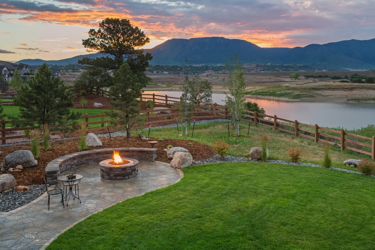 A backyard outdoor patio with a stone fire pit, curved seating, green lawn, and a view of a lake and mountains at sunset.