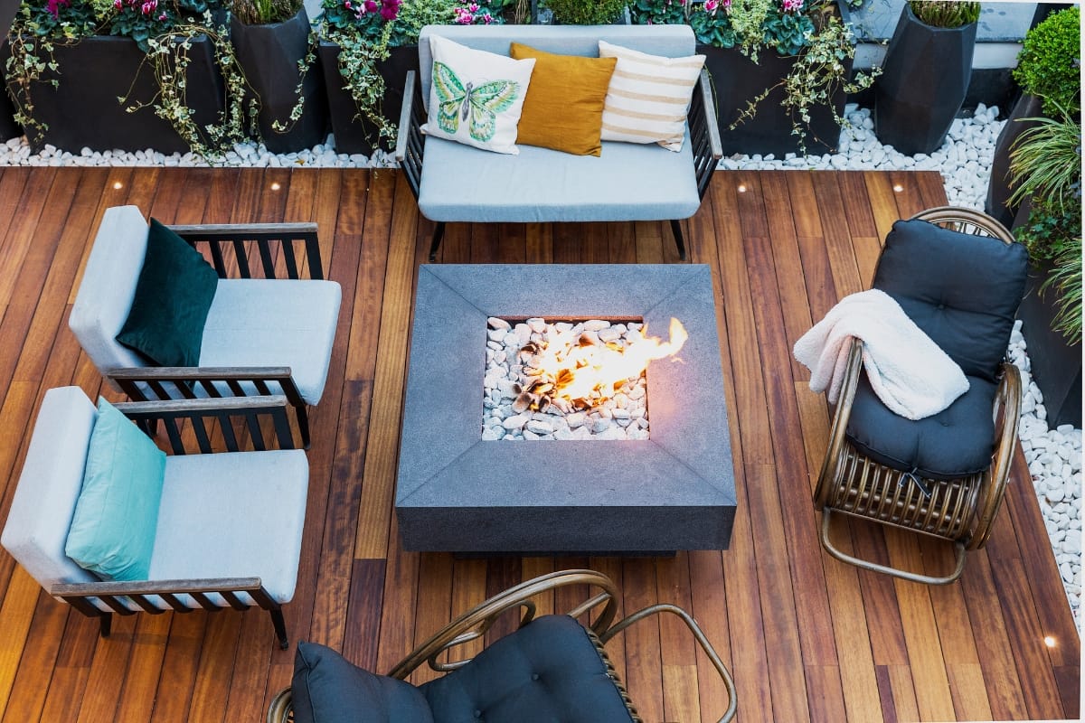 Outdoor Patios, Decks, and Fire Pits in Sandy Plains: Expert Backyard Remodeling 5 A modern outdoor patio with wooden flooring, a central square fire pit, cushioned seating, throw pillows, and decorative plants along the edges—perfect for those who appreciate stylish outdoor patios.