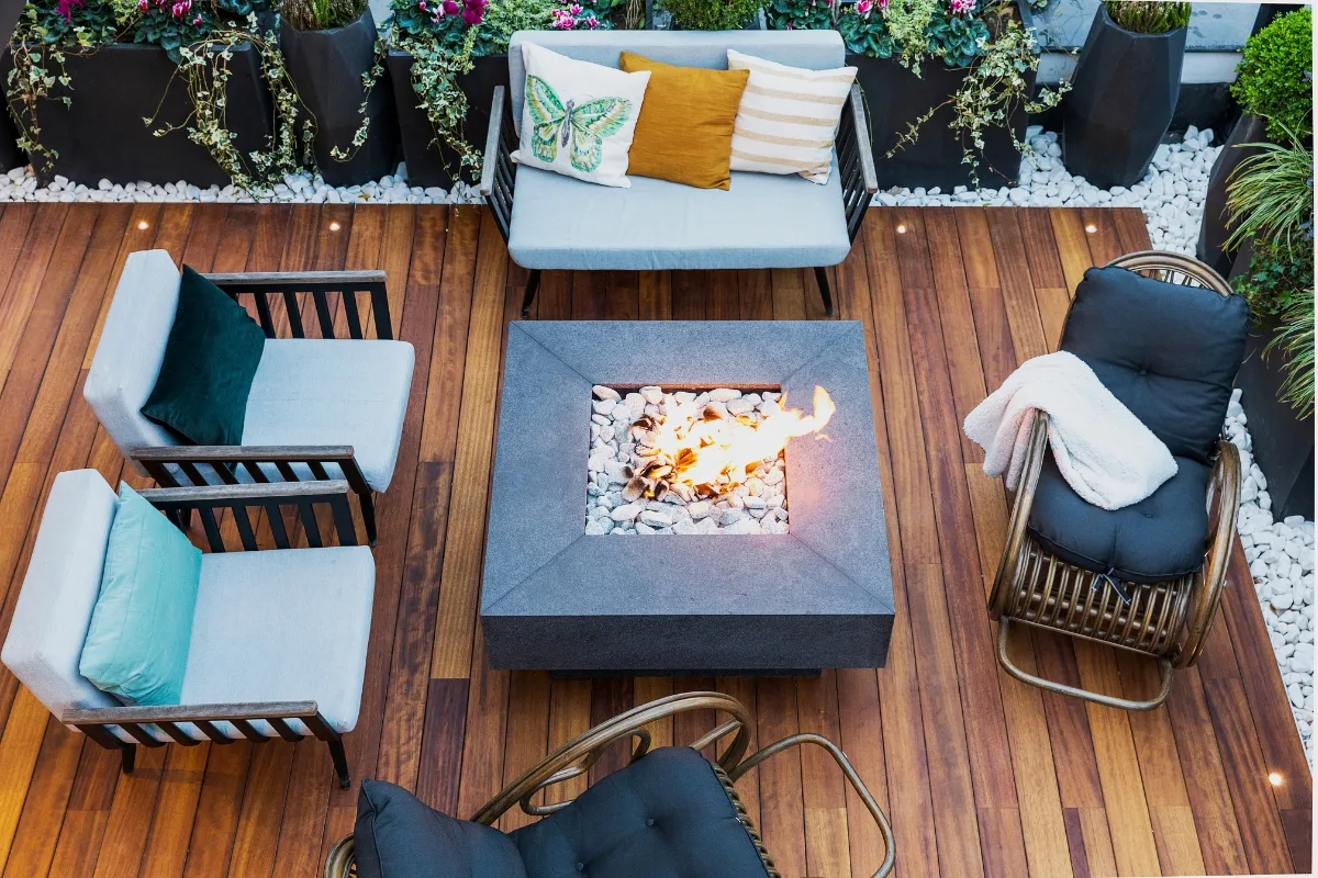 A modern outdoor patio with wooden flooring, a central square fire pit, cushioned seating, throw pillows, and decorative plants along the edges—perfect for those who appreciate stylish outdoor patios.