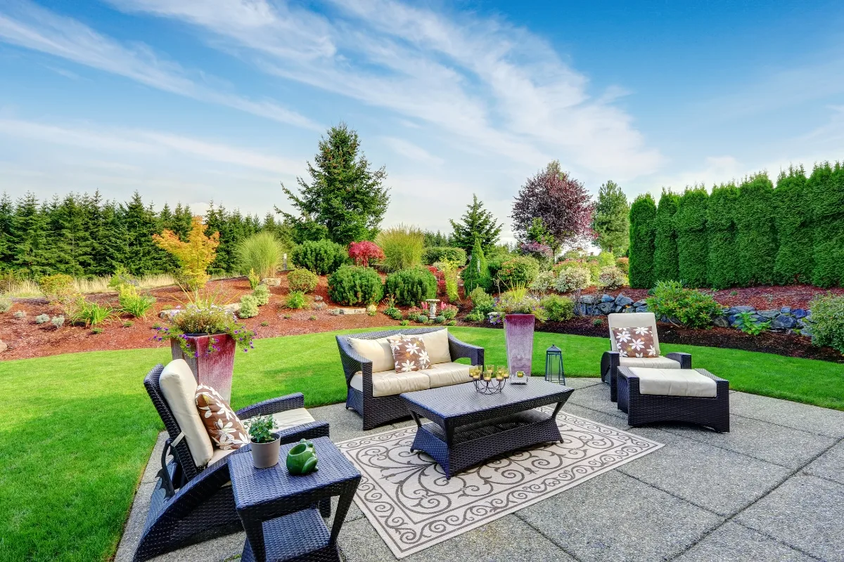 A patio with cushioned wicker furniture and a patterned rug showcases outdoor patios living, overlooking a landscaped backyard with green grass, trees, shrubs, and a blue sky.