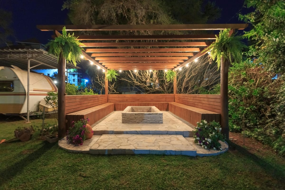 Outdoor Patios, Decks, and Fire Pits in Sandy Plains: Expert Backyard Remodeling 7 Wooden pergola with hanging plants, surrounded by benches and lit by string lights, stands on a stone patio with a built-in rectangular fire pit—creating one of the coziest outdoor patios in the garden at night.