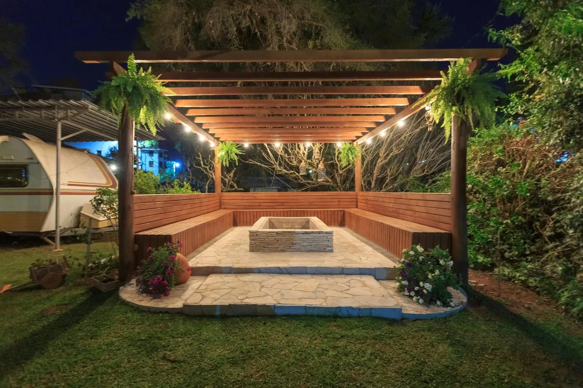 Wooden pergola with hanging plants, surrounded by benches and lit by string lights, stands on a stone patio with a built-in rectangular fire pit—creating one of the coziest outdoor patios in the garden at night.