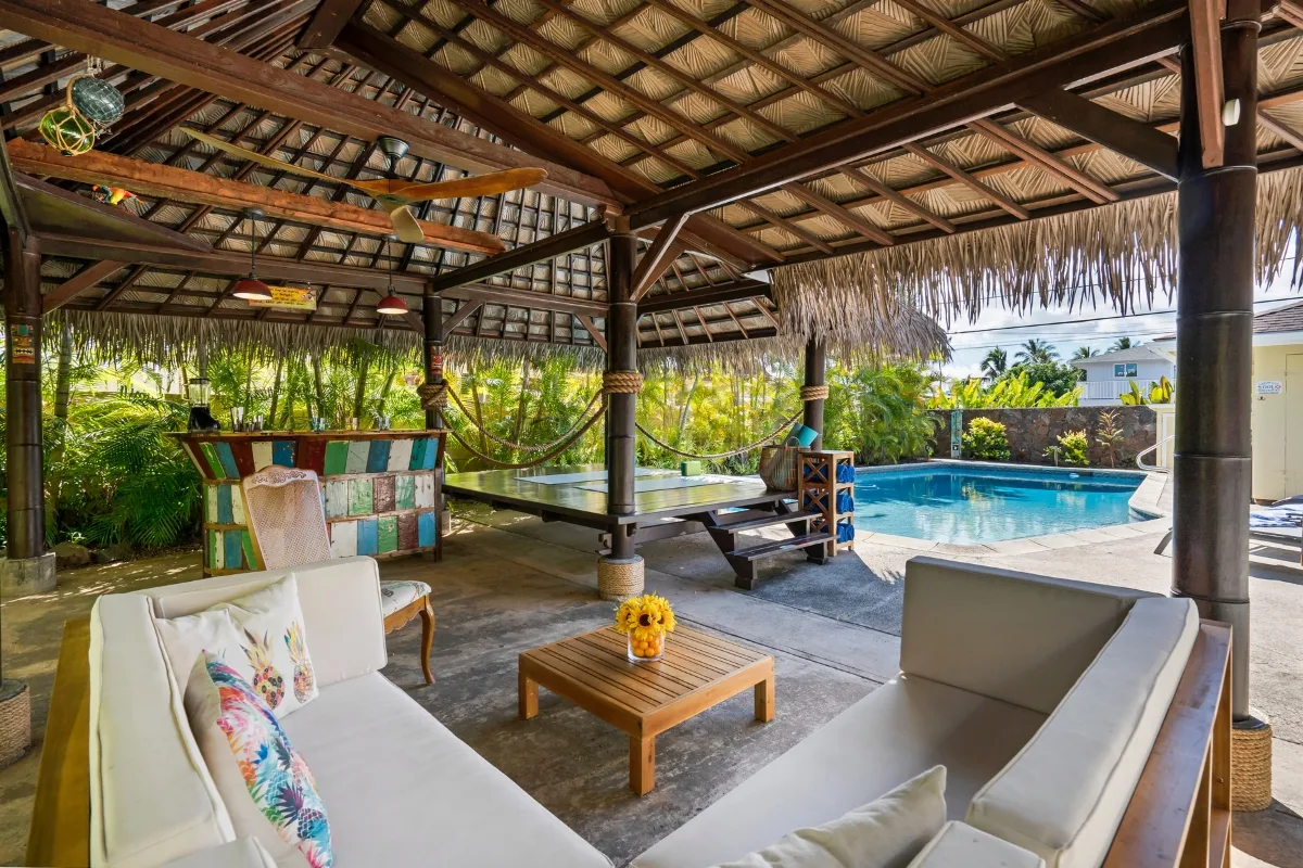 This inviting outdoor patio features a thatched roof, white sofas, a table with sunflowers, a hammock, pool table, and swimming pool all surrounded by lush tropical plants—a perfect example of stylish outdoor patios.