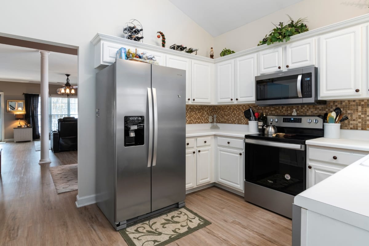 Smart Home Remodeling in Sandy Springs: Top Energy-Efficient Upgrades 5 Modern kitchen with white cabinets, stainless steel refrigerator, oven, and microwave; featuring a mosaic tile backsplash and wood flooring. Open doorway leads to a living area—perfect for smart home remodeling enthusiasts.