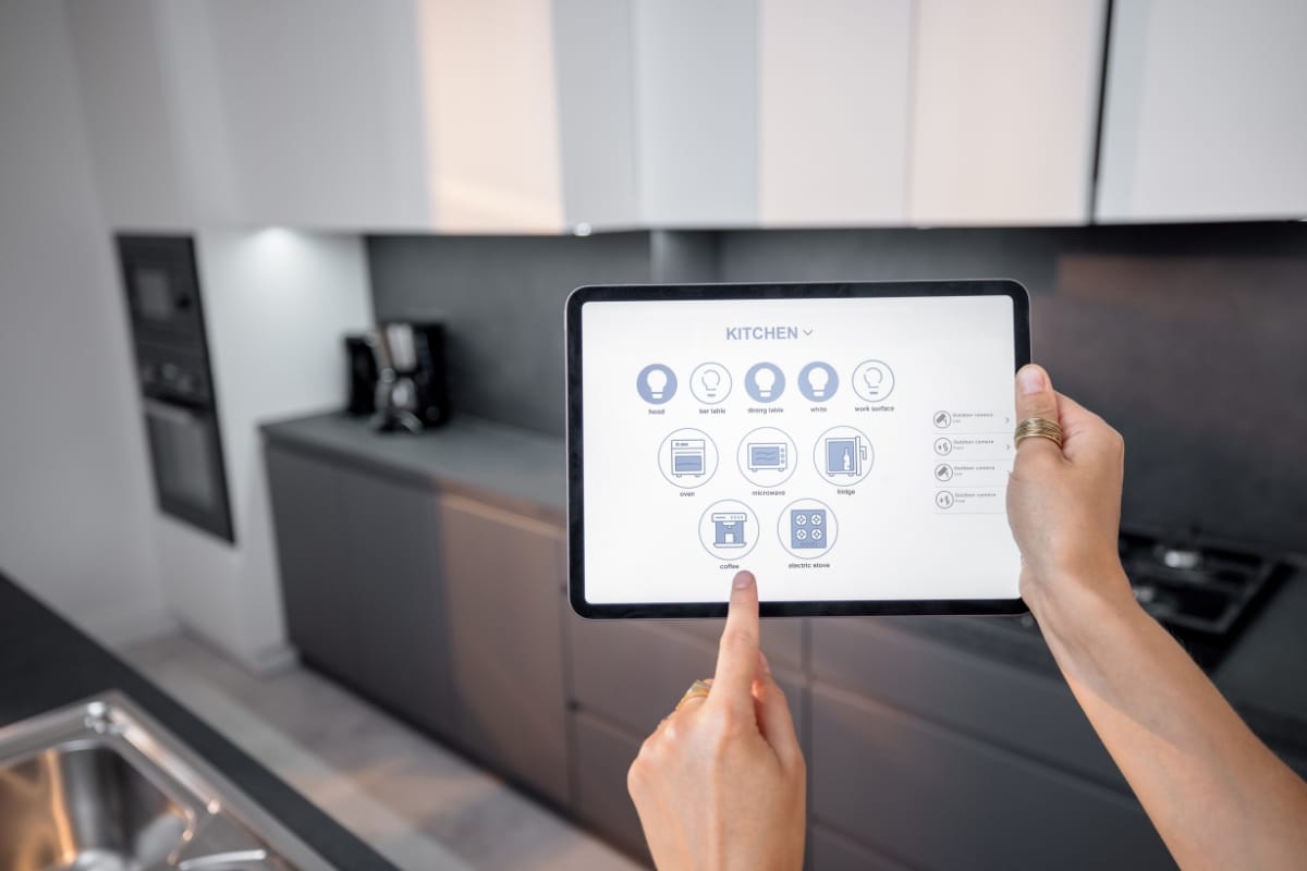 Smart Home Remodeling in Sandy Springs: Top Energy-Efficient Upgrades 7 Person uses a tablet to control smart kitchen devices, showcasing various appliance icons on the screen; a modern kitchen designed with smart home remodeling is visible in the background.
