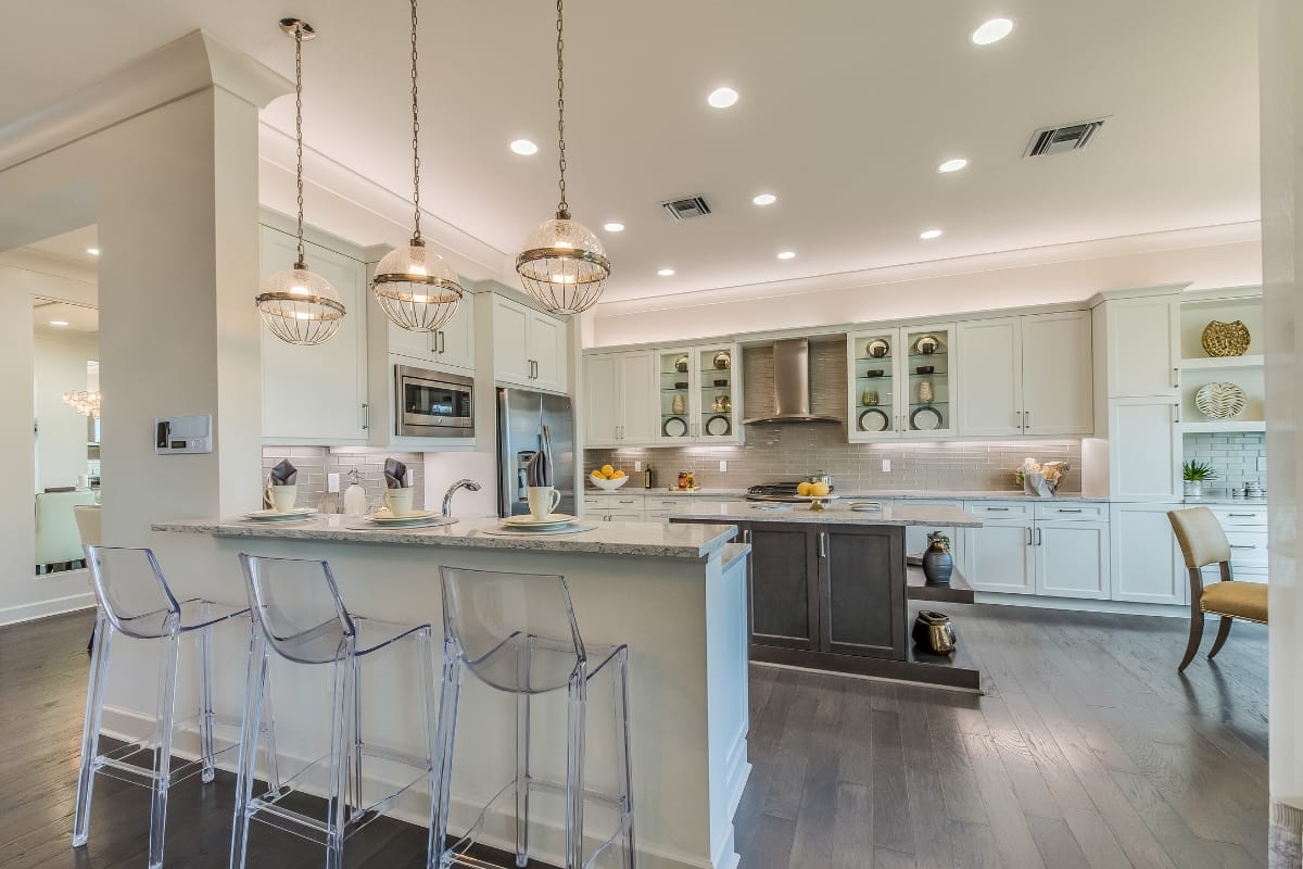 Modern kitchen with white cabinets, pendant lights, a center island, clear bar stools, stainless steel appliances, and dark wood flooring—perfect for those seeking affordable kitchen upgrades.