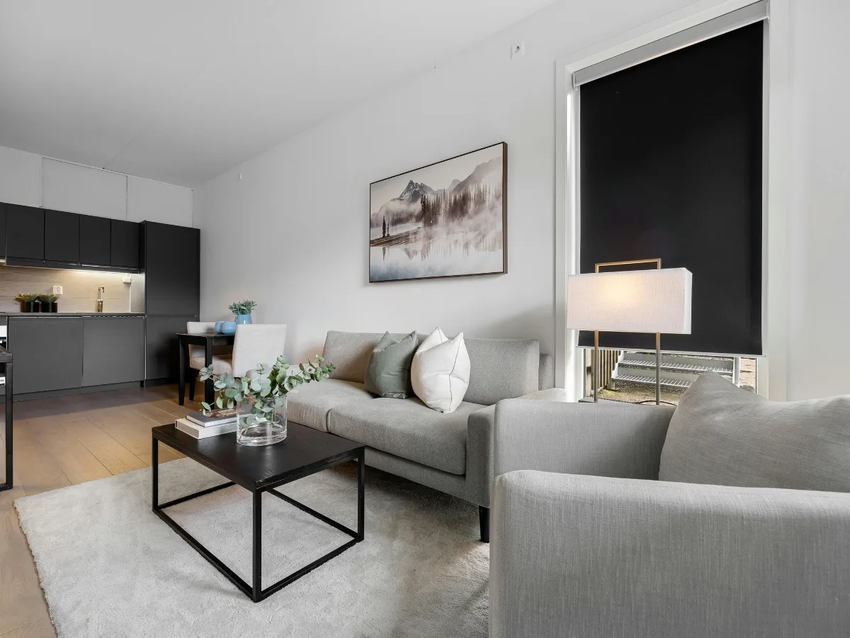 Modern living room with light gray sofas, a black coffee table, a floor lamp, and a large window with a black shade; kitchen and dining area visible in the background&mdash;perfect for stylish condo remodeling.