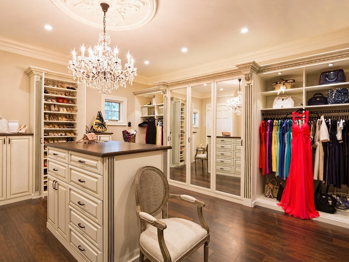 Spacious walk-in closet designed by custom closet designers, featuring chandeliers, an island with drawers, shelves for shoes and handbags, hanging clothes, a window, and a cushioned chair on dark wood flooring.